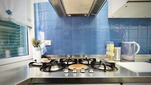 A kitchen with a gas stove, blue tiles, a window with blinds, and kitchen utensils. - Splendida Dimora Negri (Genoa)