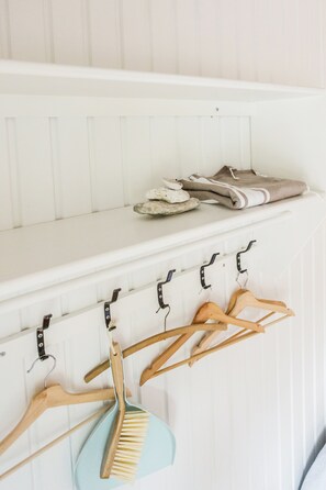 A white wall with a white shelf holding folded clothes and a blue brush hanging below a coat rack.