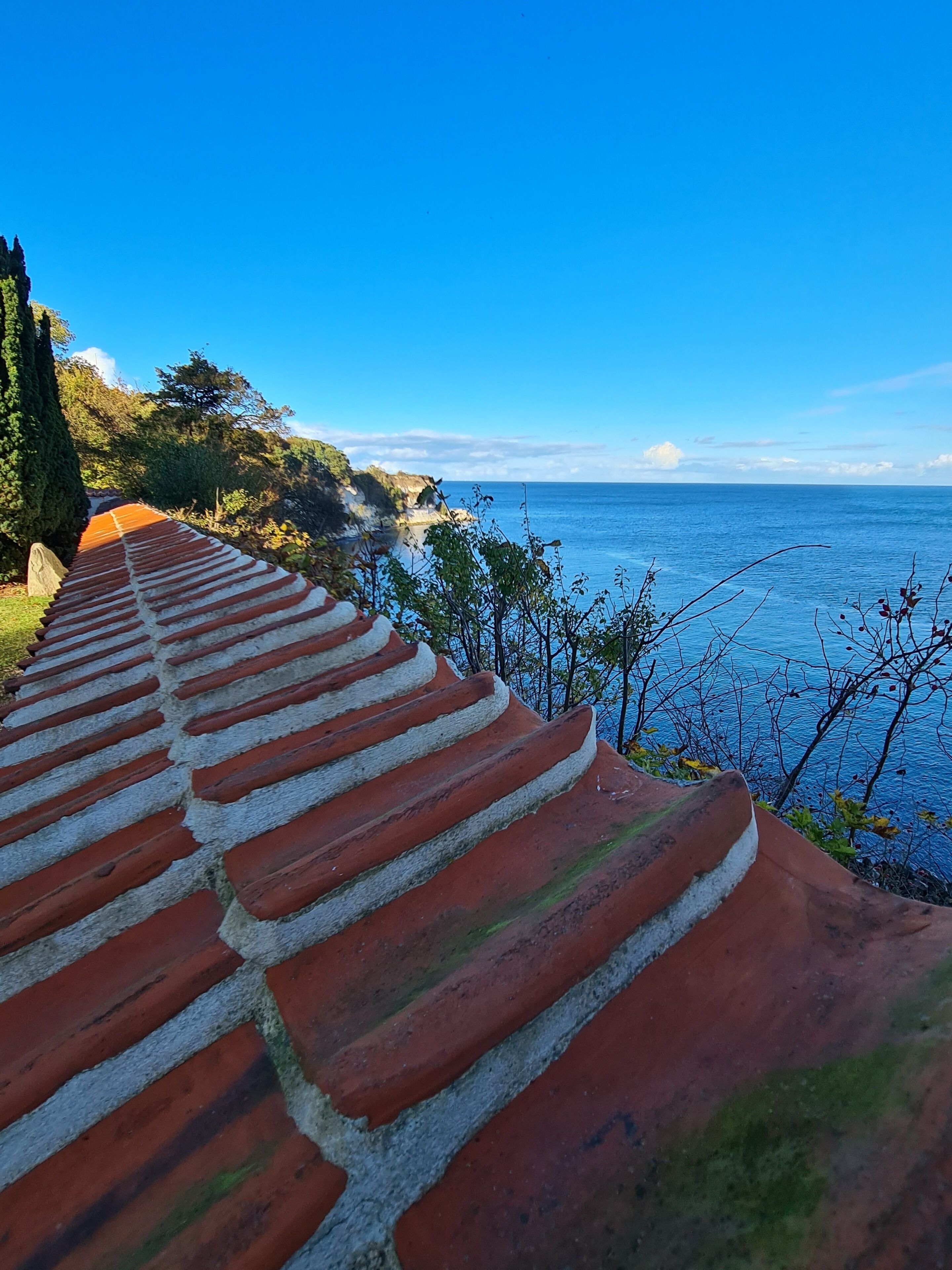 A coastal pathway with a red and white tiled roof, leading to a beach with clear blue water and a rocky shoreline.