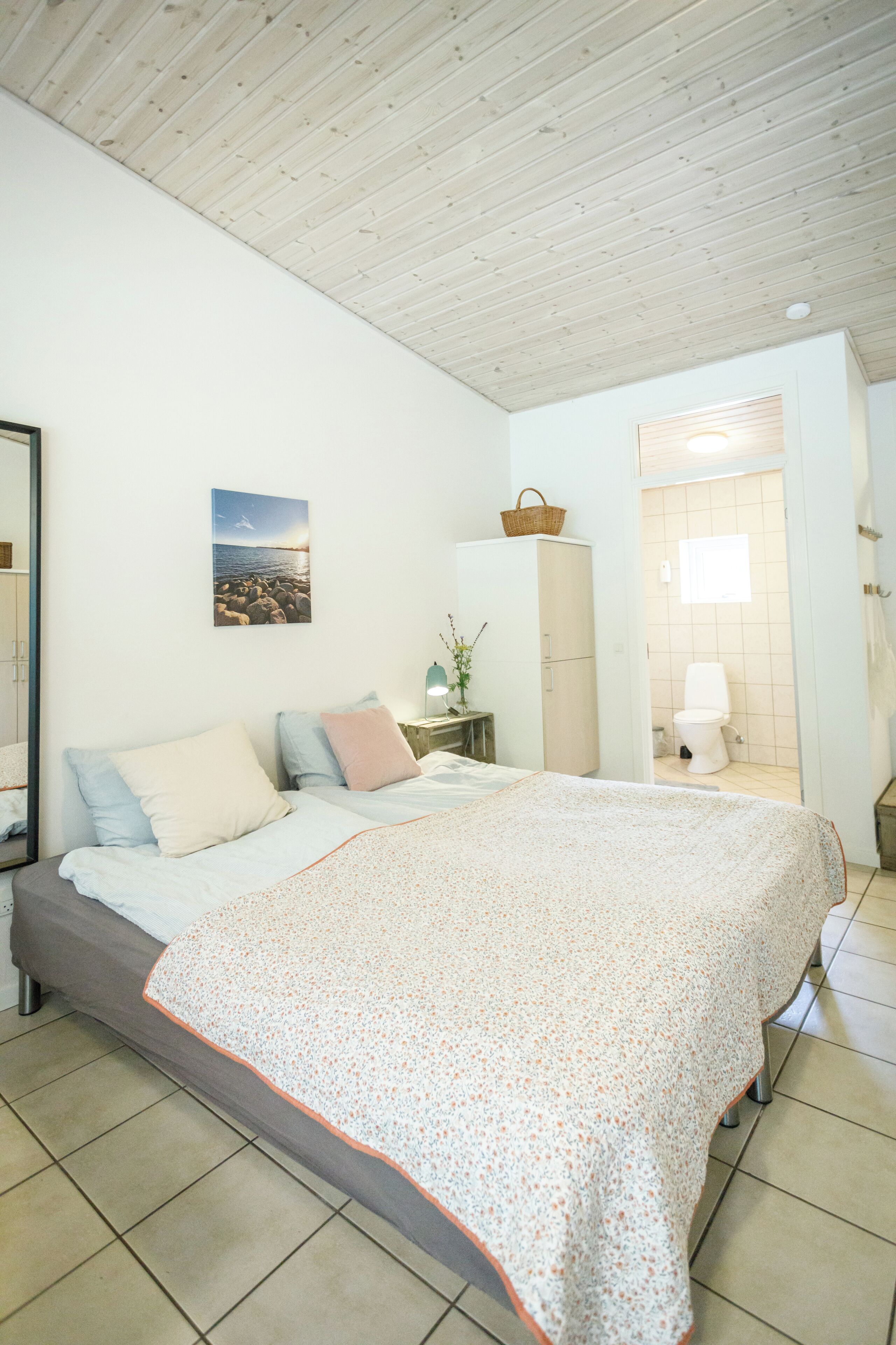 Un dormitorio con cama, almohadas, una mesita de noche, una lámpara, un cuadro en la pared y un baño visible a través de una puerta abierta.