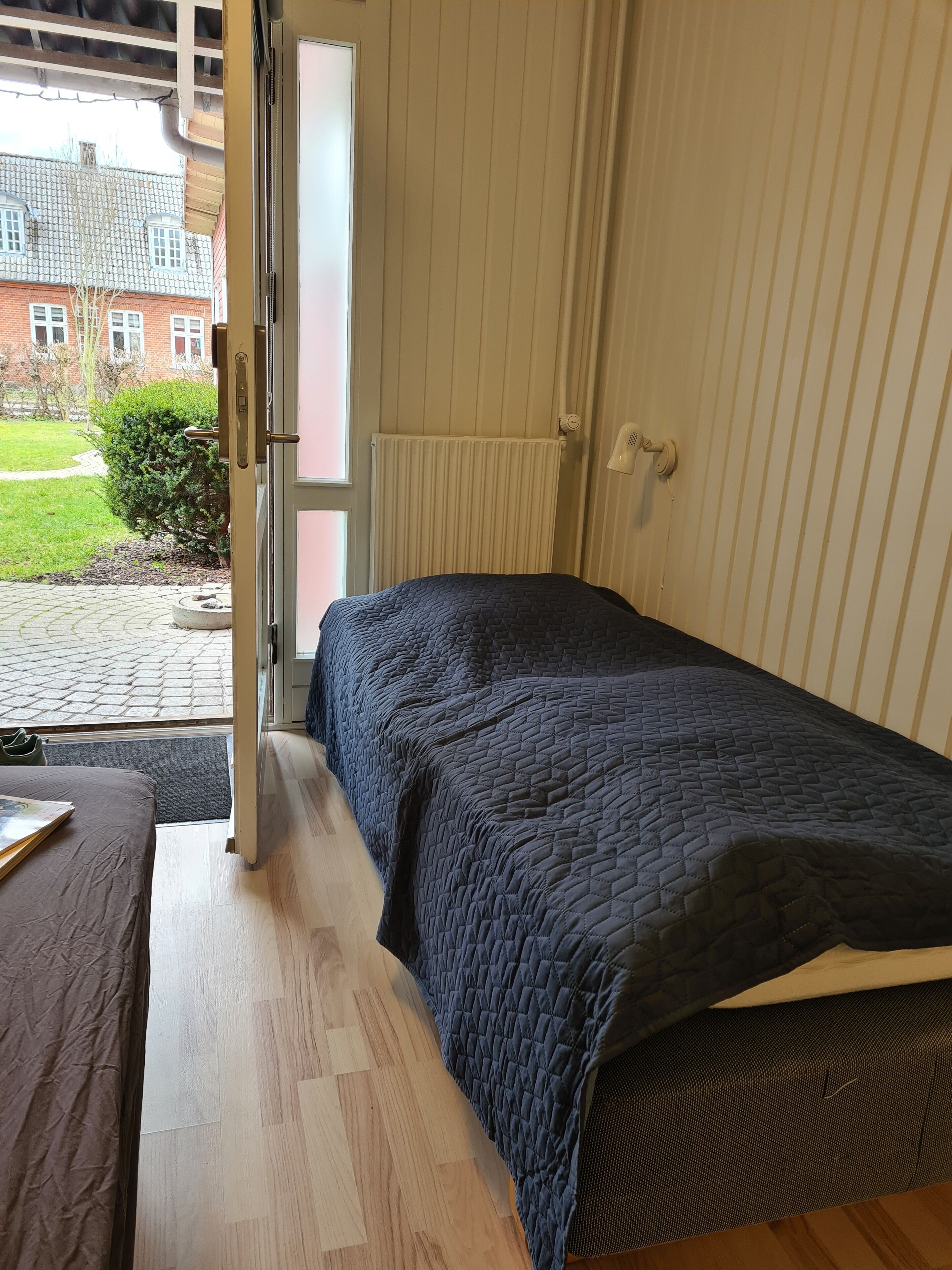 Ein Schlafzimmer mit Bett, Nachttisch und Blick auf ein Backsteinhaus sowie Grünflächen.
