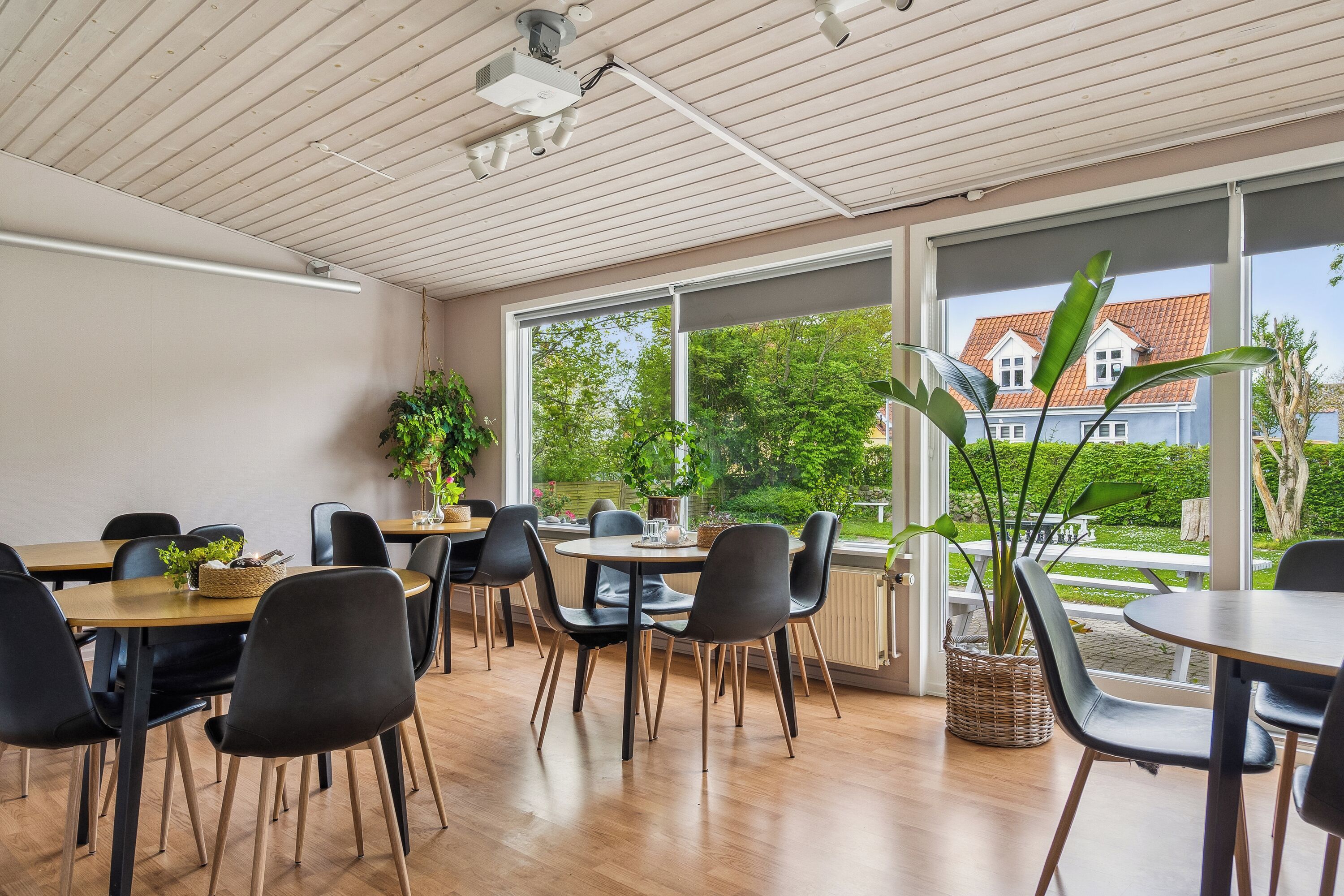 A dining area with wooden tables and black chairs, large windows with a view of a house and greenery, and ceiling-mounted lights.
