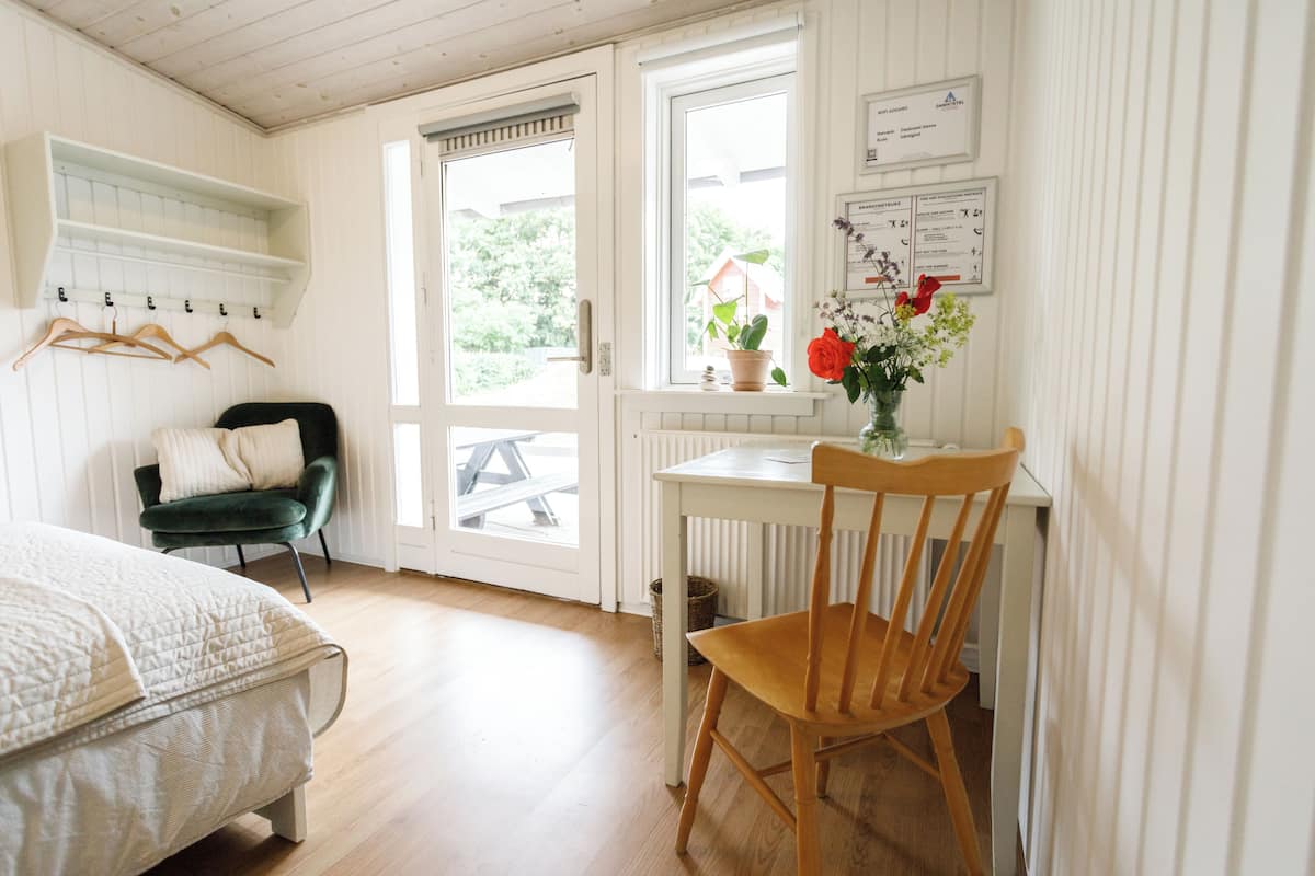 Ett rum med en säng, ett skrivbord med en stol, ett fönster med utsikt över grönområden och en hylla med galgar.