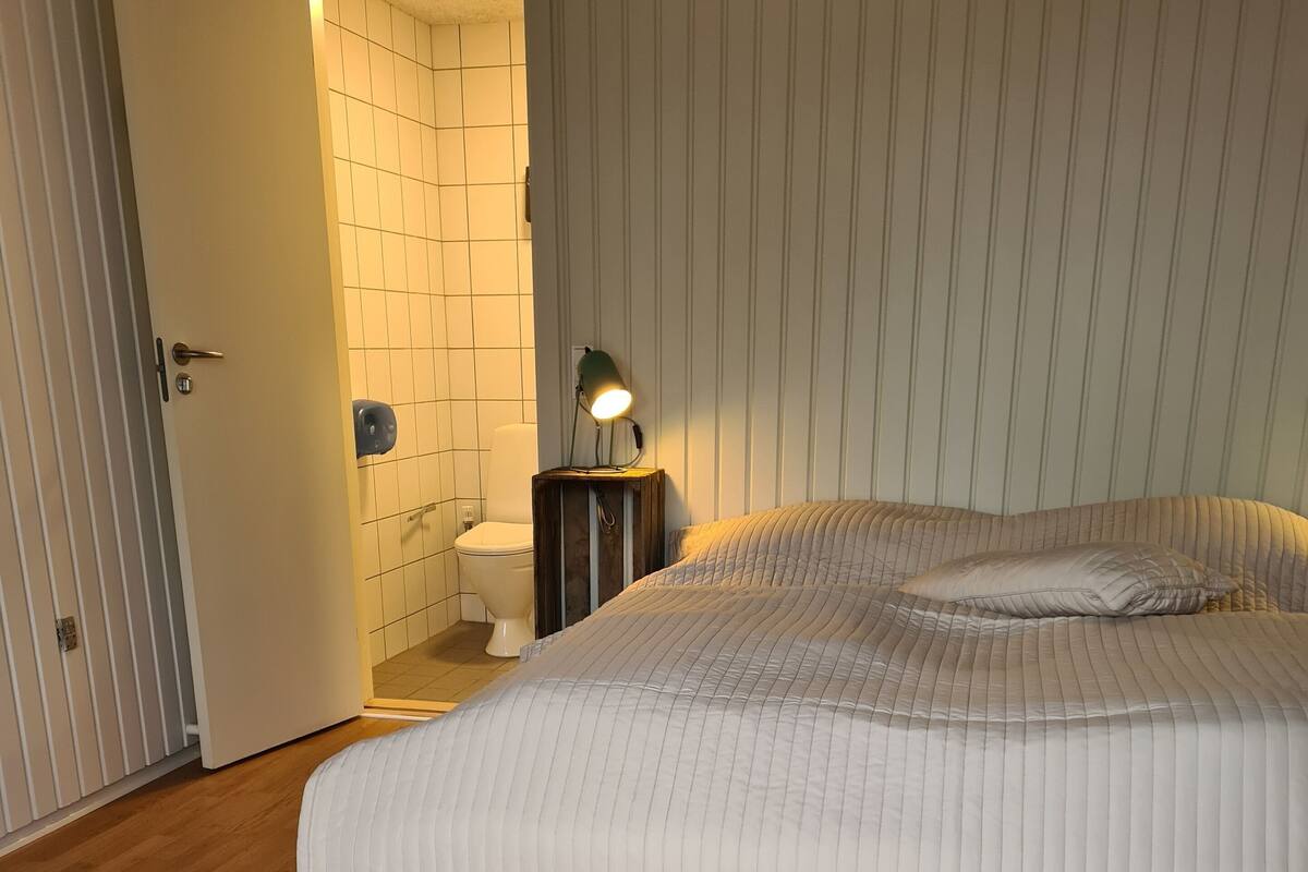 Ett sovrum med trägolv, en säng med ett quiltat överkast, ett sängbord med en lampa och ett badrum med toalett och handtork.