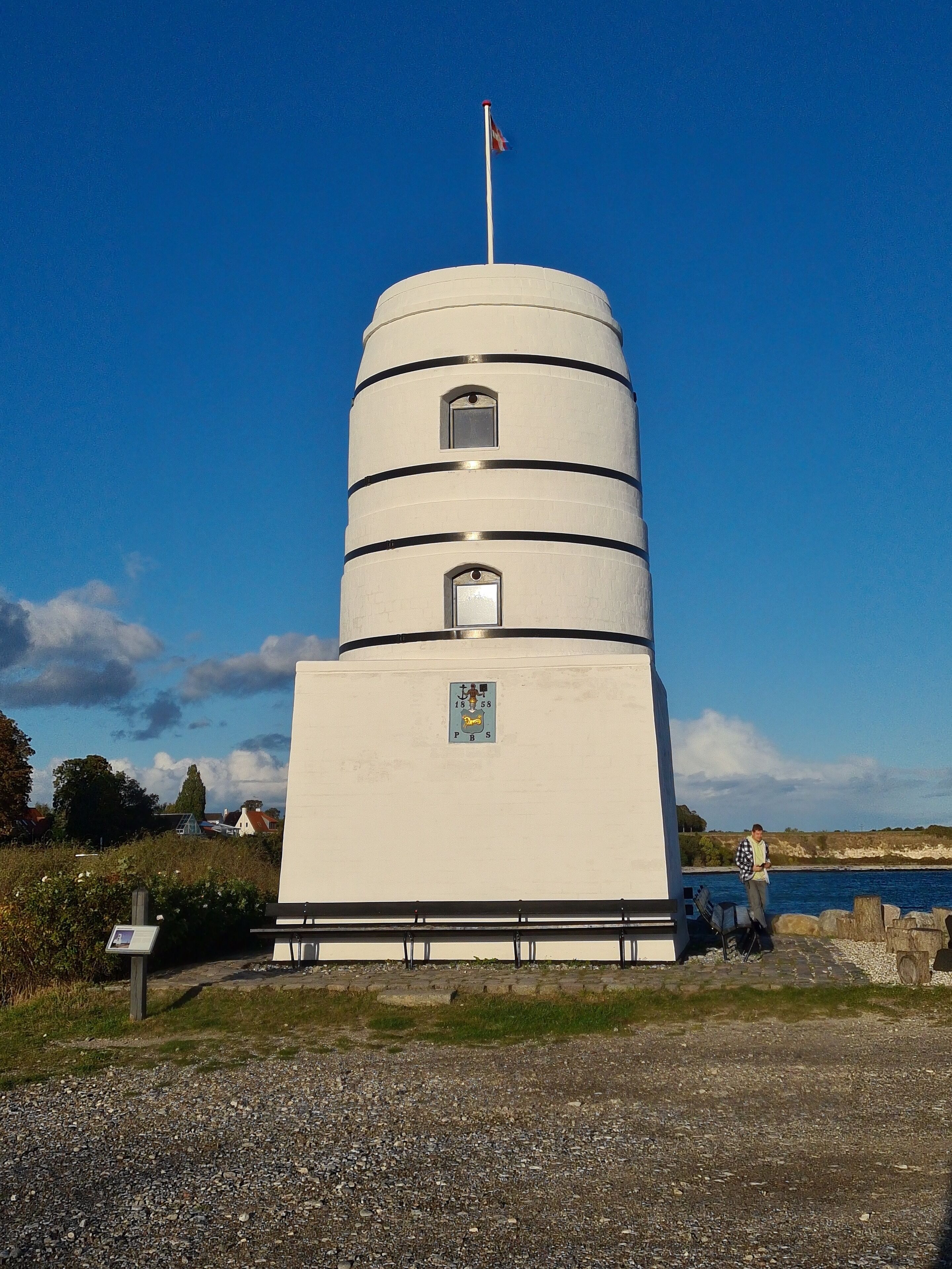 A cylindrical white building with a flag on top, a small window, and a plaque with text and a coat of arms.