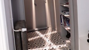 Ein schmaler Flur mit einem gemusterten Boden, einer Tür am Ende und einem Bücherregal auf der rechten Seite.