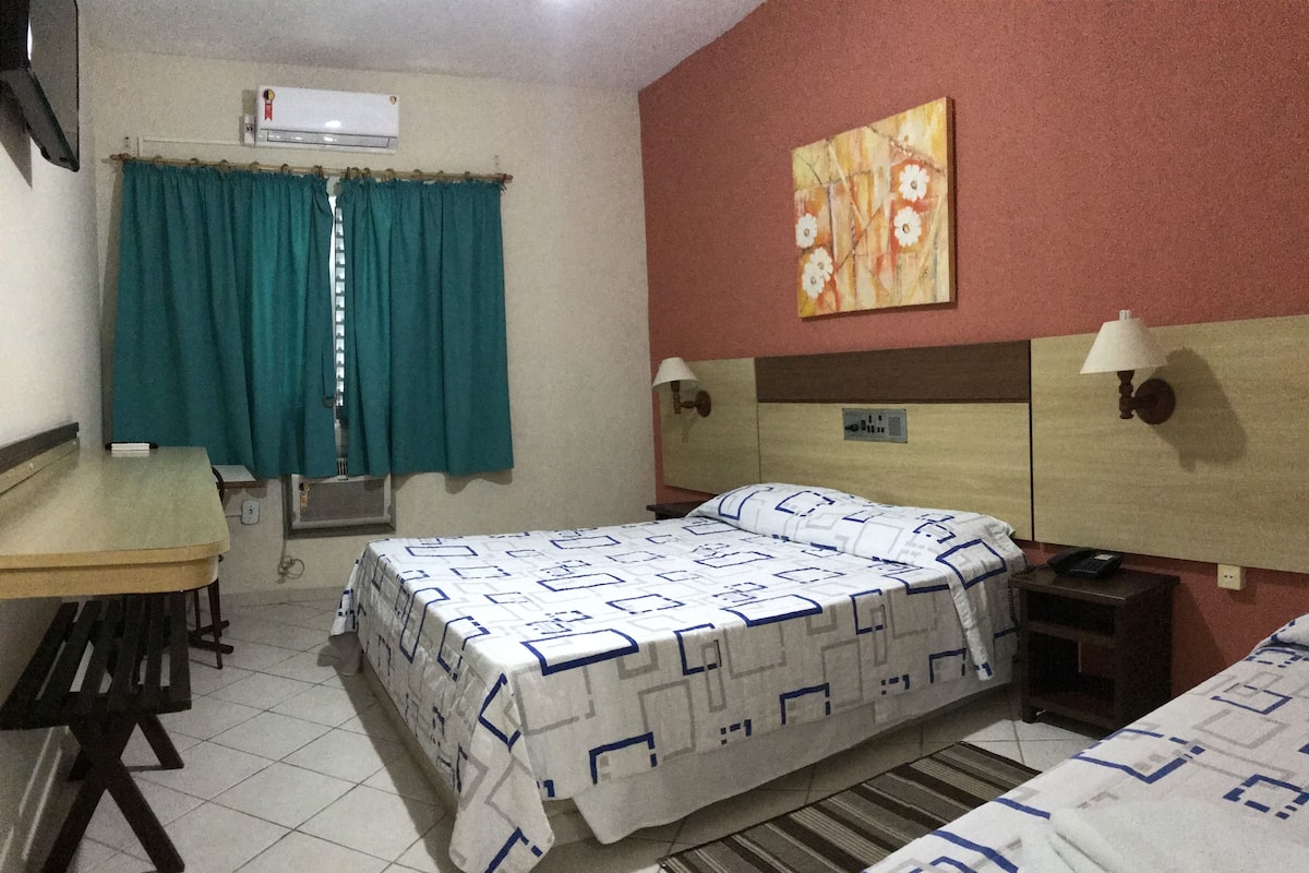A hotel room with a bed, a desk, and a chair. There is a window with curtains, a wall-mounted air conditioner, and a painting on the wall.