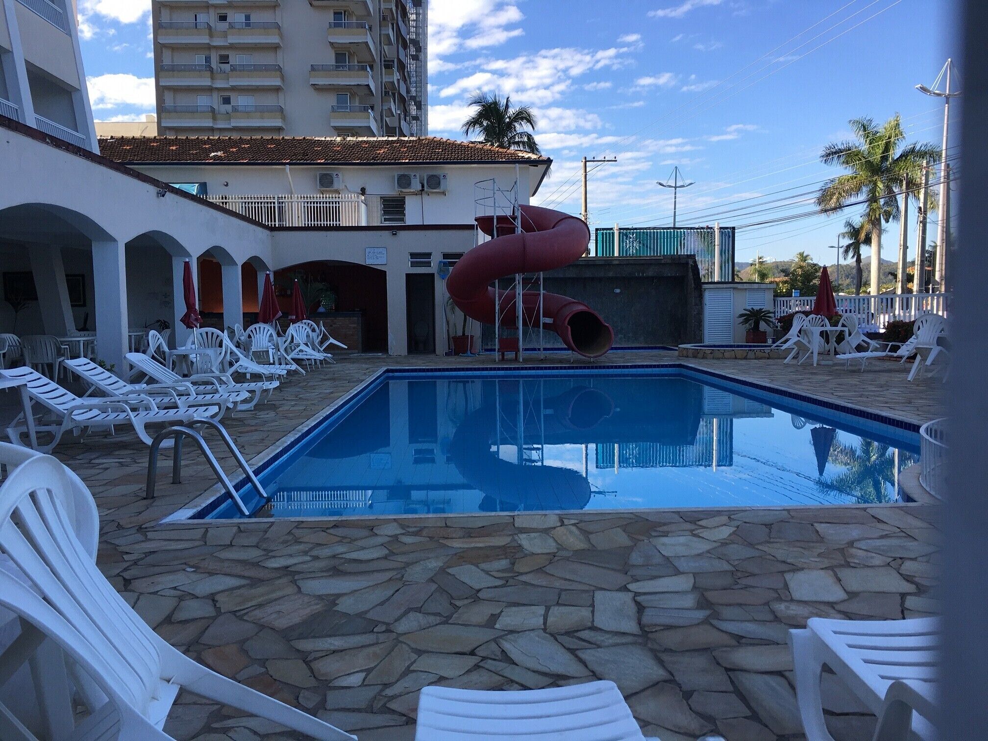 Une piscine avec un toboggan rouge, entourée de chaises longues blanches et d’une terrasse dallée de pierres.