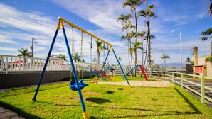 A playground with swings and slides, surrounded by a white fence and palm trees.