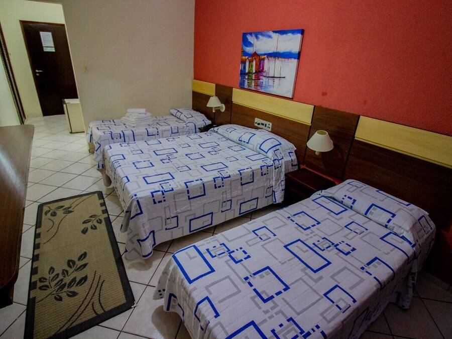 A hotel room with three beds, a wooden desk, a red wall with a painting, and a tiled floor.