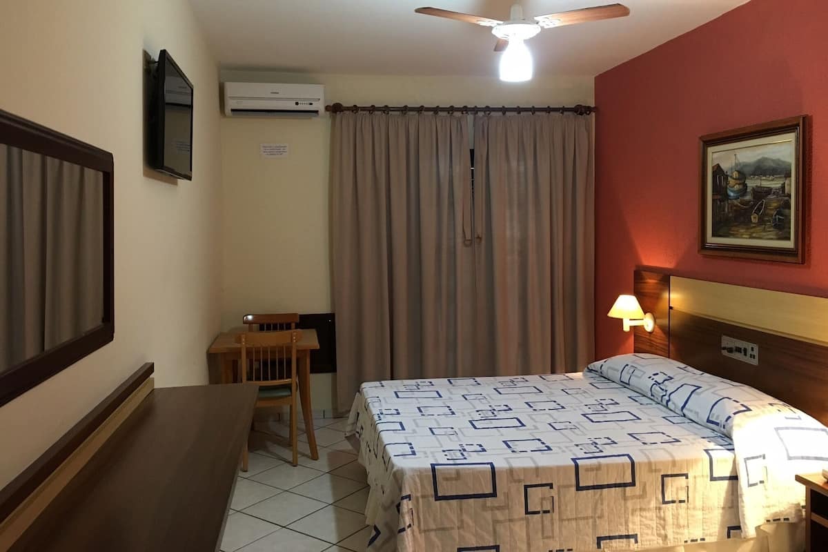 A hotel room with a bed, a desk, a chair, a ceiling fan, an air conditioner, a lamp, and a painting on the wall.