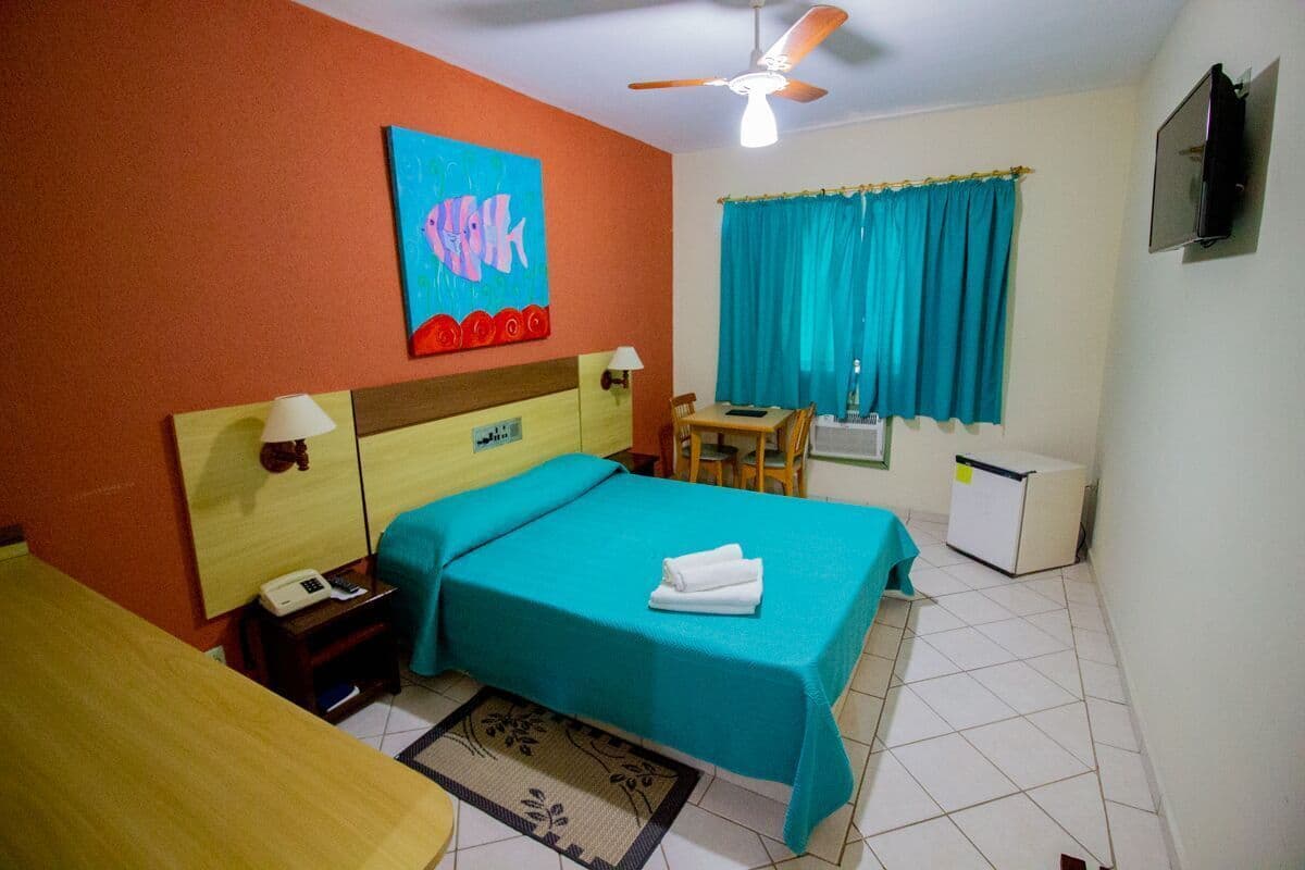 A hotel room with a bed, a desk, a chair, a small refrigerator, a ceiling fan, and a painting on the wall.