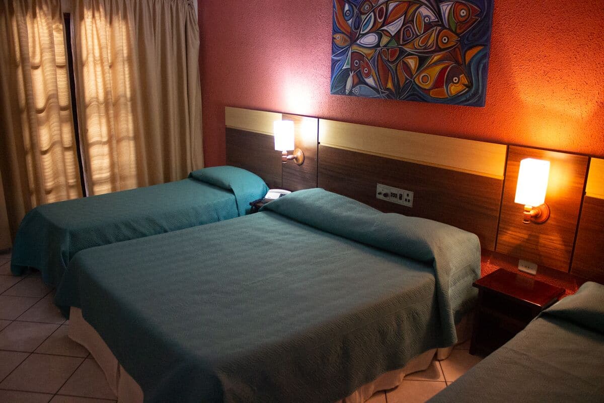 A hotel room with two beds, a headboard with wall-mounted lights, a painting on the wall, and a window with curtains.