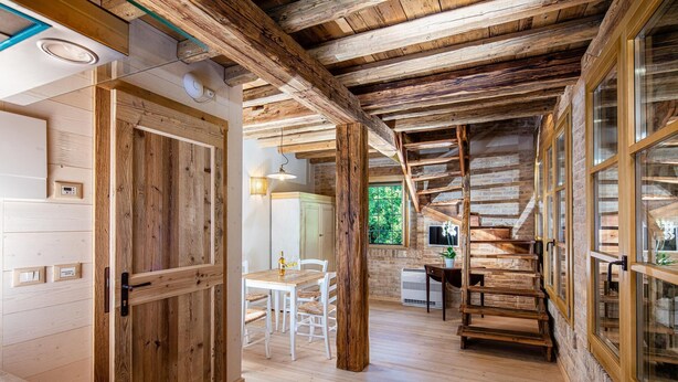 Un interior rústico de madera con vigas a la vista, una zona de comedor con sillas blancas y una escalera.