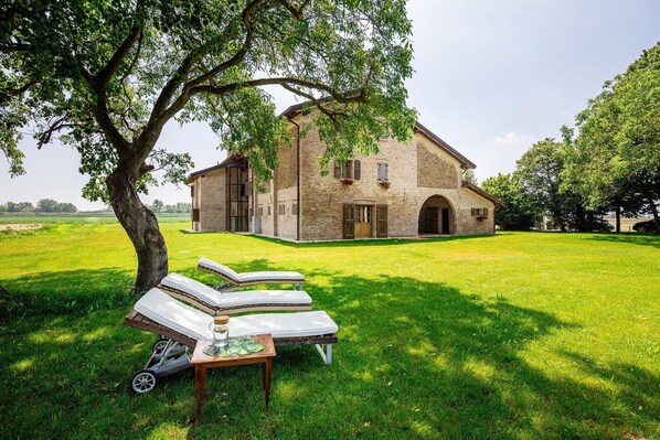 A rustic brick building with a large lawn, two lounge chairs, and a table with a pitcher and glasses. - Agriturismo Casa Gioia (San Michele al Tagliamento)