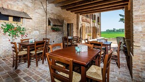 Un área de comedor al aire libre de estilo rústico, con paredes de ladrillo, mesas y sillas de madera, y vistas a un campo verde.