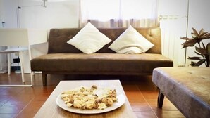 Una sala de estar con un sofá marrón, dos cojines blancos, una mesa de centro blanca con un plato de nueces y una planta en la esquina.