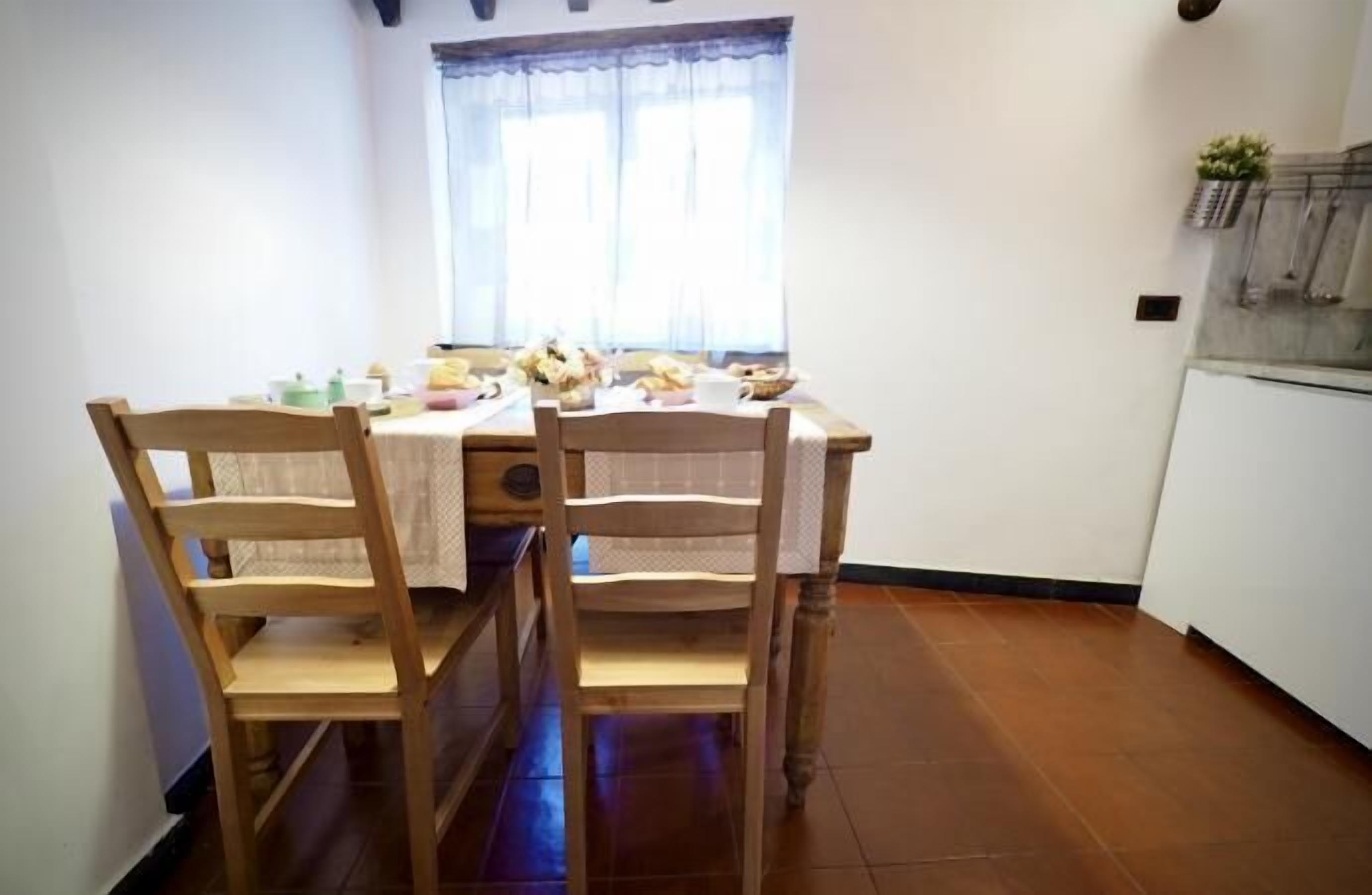 A dining area with a wooden table and chairs, a window with curtains, and a kitchen area with a refrigerator.