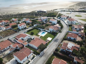 Vue aérienne d’un quartier résidentiel situé sur la côte, avec des maisons dotées de toits recouverts de tuiles rouges, une piscine centrale et une plage en arrière-plan.
