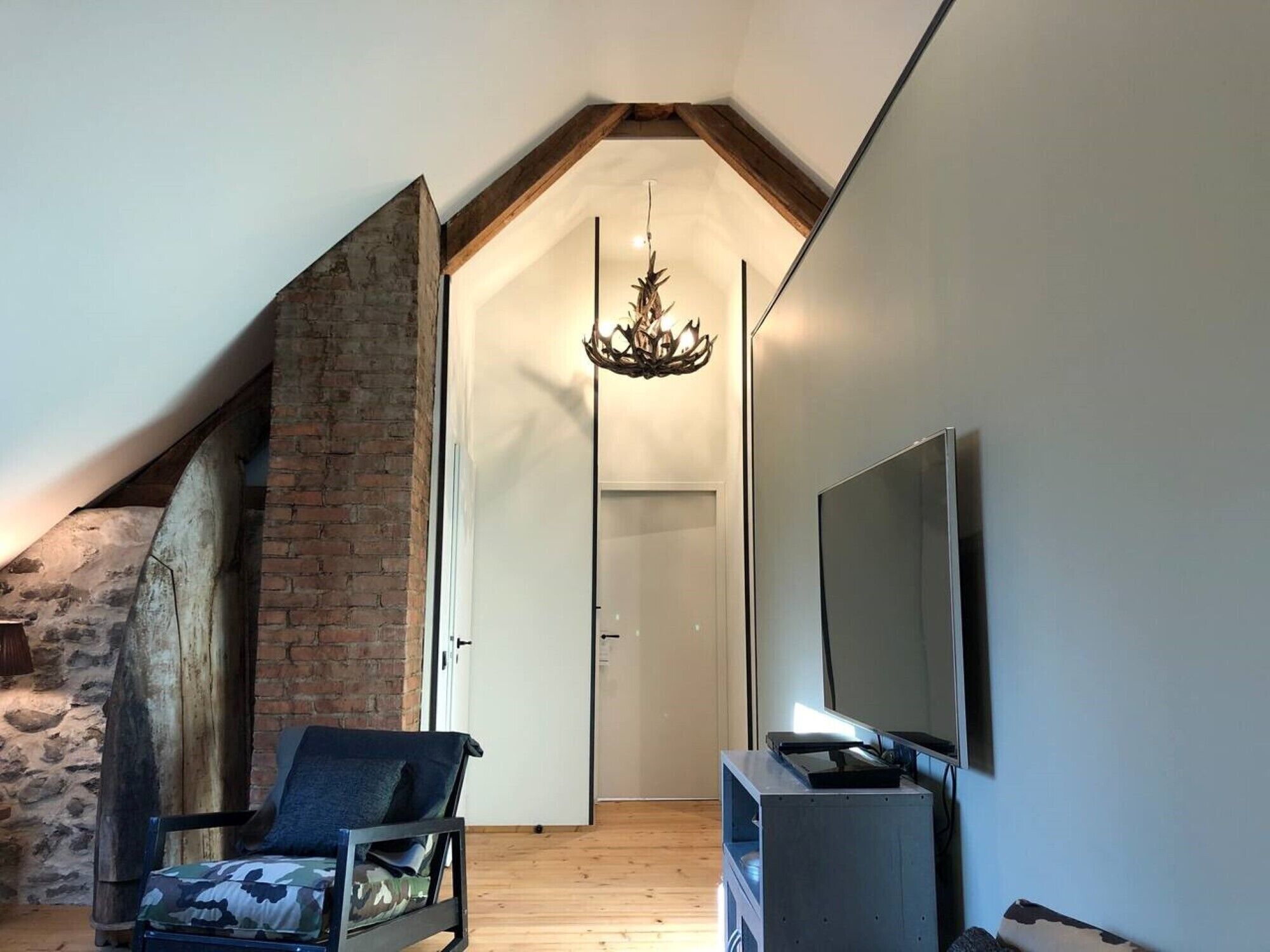 A loft-style room with a sloped ceiling, exposed brick wall, a wooden staircase, a modern chandelier, a television, and a comfortable chair.