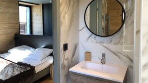 Une chambre à coucher moderne avec un lit, une tête de lit en bois, une grande fenêtre et une salle de bain avec un mur en marbre, un miroir rond et un lavabo.