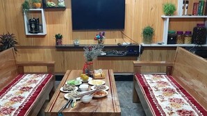 A room with wooden walls and a central table set with various dishes. There are two wooden benches with red and white patterned cushions. A shelf with books and decorative items is visible on the wall. - Homestay A San - Hostel (Meo Vac)