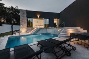 Ein moderner Poolbereich im Freien mit einer Steinmauer, einer eingelassenen Sitzbank und einer Treppe, die zu einer überdachten Terrasse führt.