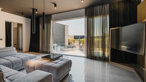 Un salon moderne avec un canapé gris, un téléviseur à écran plat et une porte vitrée donnant sur une terrasse extérieure.