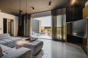 A modern living room with a grey sofa, a flat-screen TV, and a glass door leading to an outdoor patio. - Modern 3-BR villa for 6 with pool, surrounded by pine forest near beach (Valica)