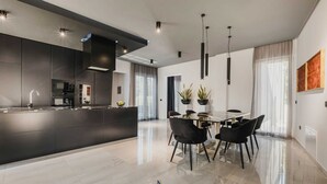 Une cuisine moderne dotée d’un îlot central, d’un coin repas avec une table en verre et des chaises noires, et d’une grande fenêtre agrémentée de rideaux légers.