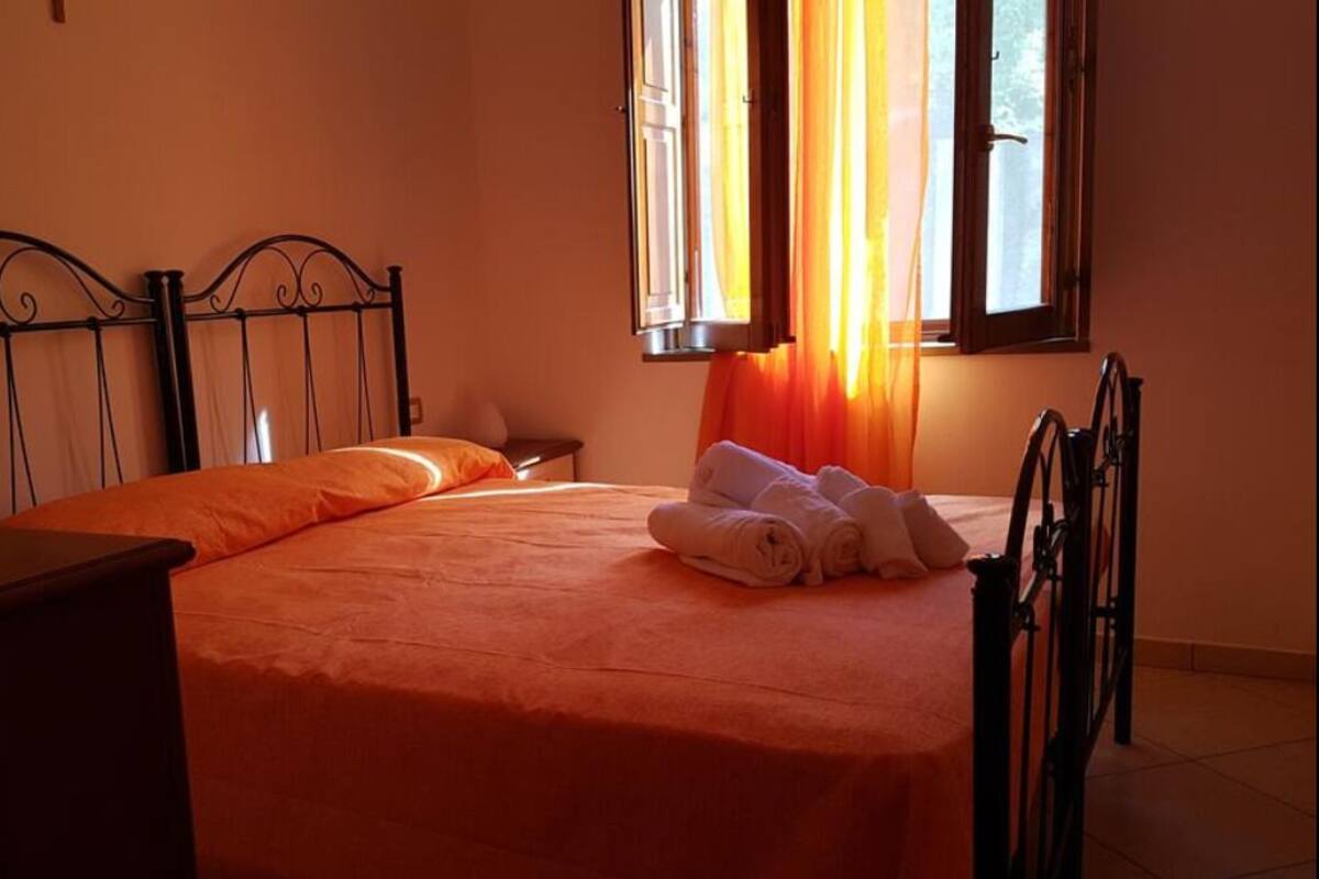Ein Einzelbett mit orangefarbener Bettdecke, ein Nachttisch aus Holz, ein Stuhl und ein Fenster mit Vorhängen.