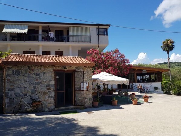 A two-story building with a stone facade and a tiled roof. There is a balcony with a white awning, and a small outdoor seating area with umbrellas and potted plants. - Il Moro della Molpa (Centola)