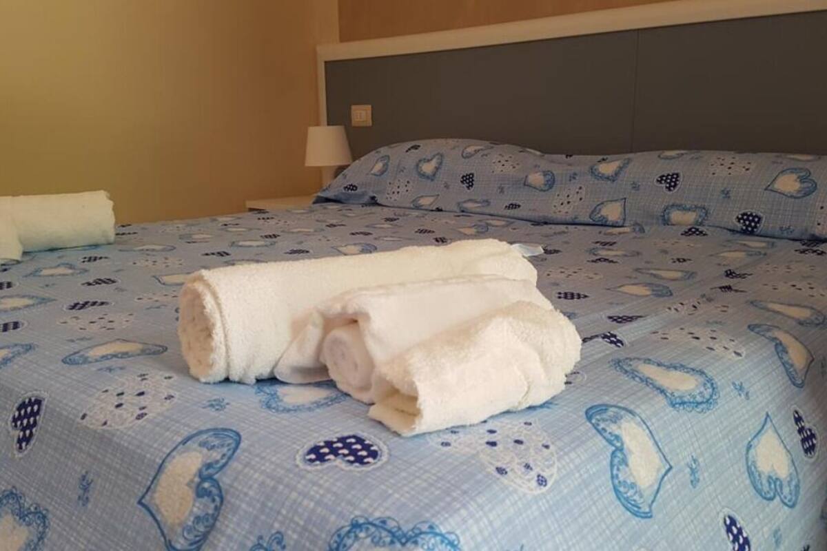 Ein ordentlich gemachtes Bett mit einer blau-weißen Bettdecke, zwei aufgerollten weißen Handtüchern und einer Nachttischlampe.