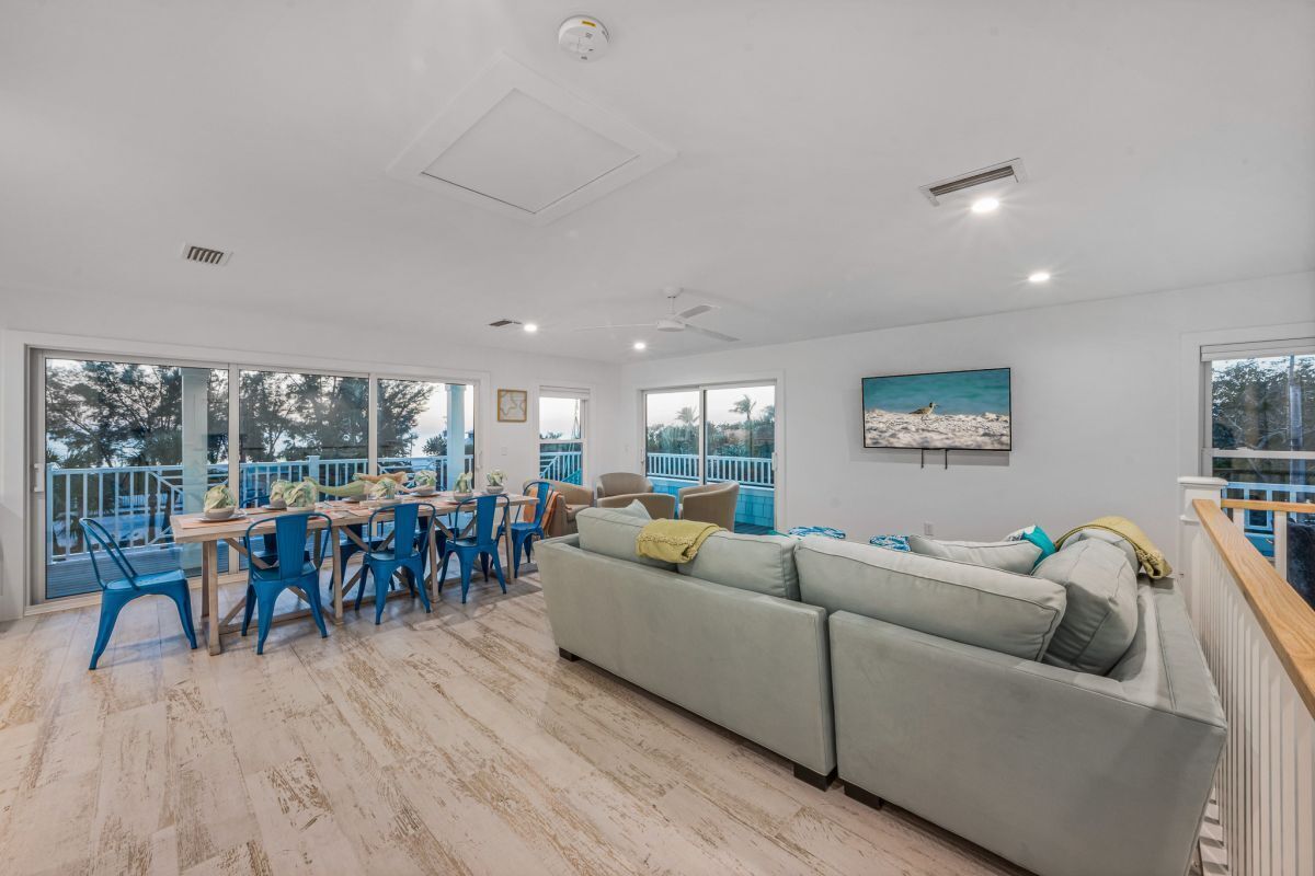 A spacious living area with a large sofa, dining table, and chairs. There are large windows offering a view of the outdoors.