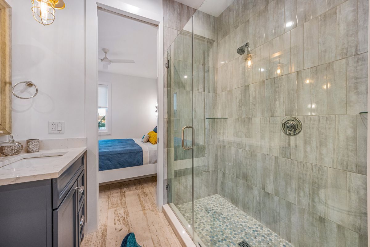 A modern bathroom with a glass shower enclosure, a white sink, and a visible bed in the adjacent room.