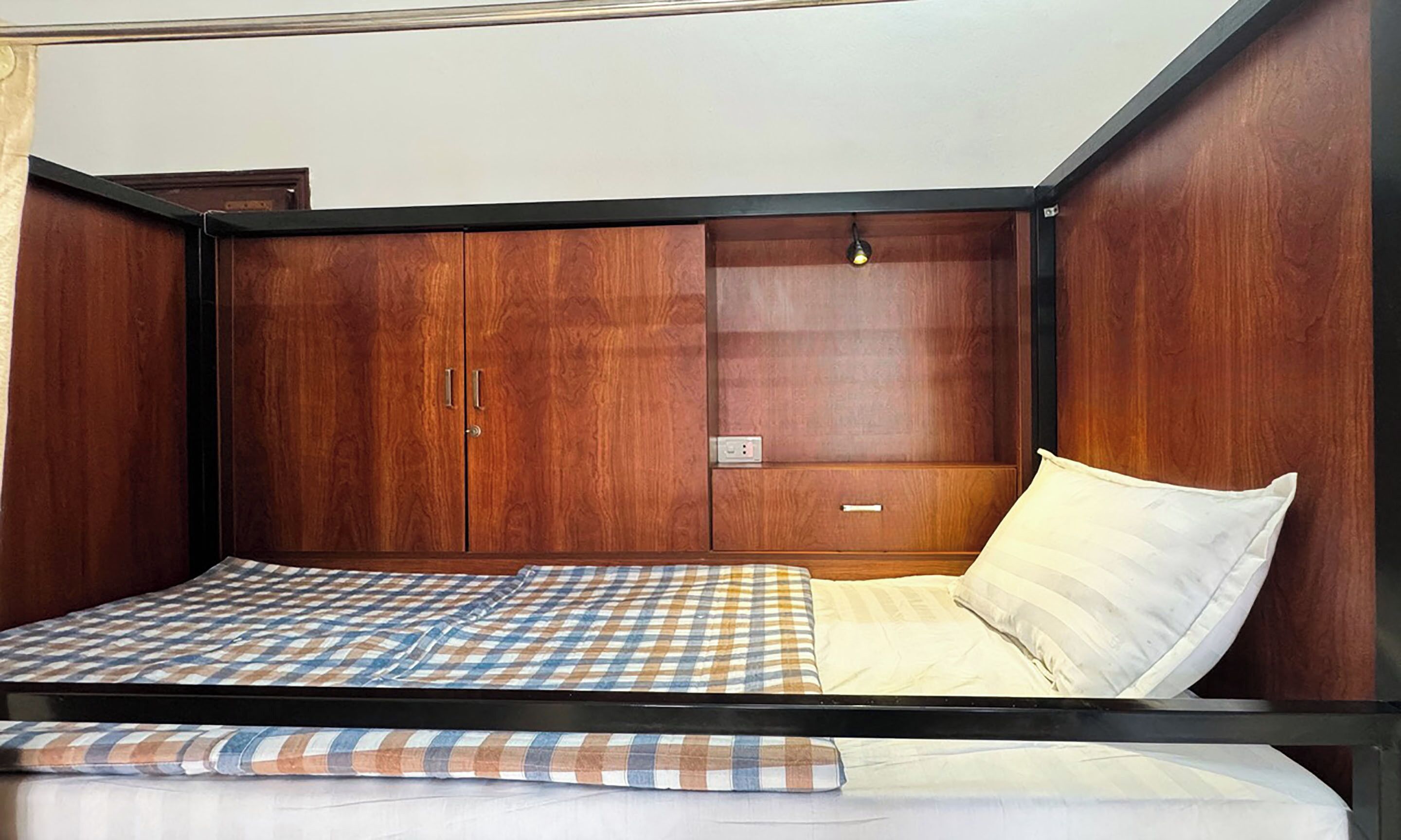 A single bed with a checkered bedspread, a wooden wardrobe with a mirror, and a small bedside table with a lamp.