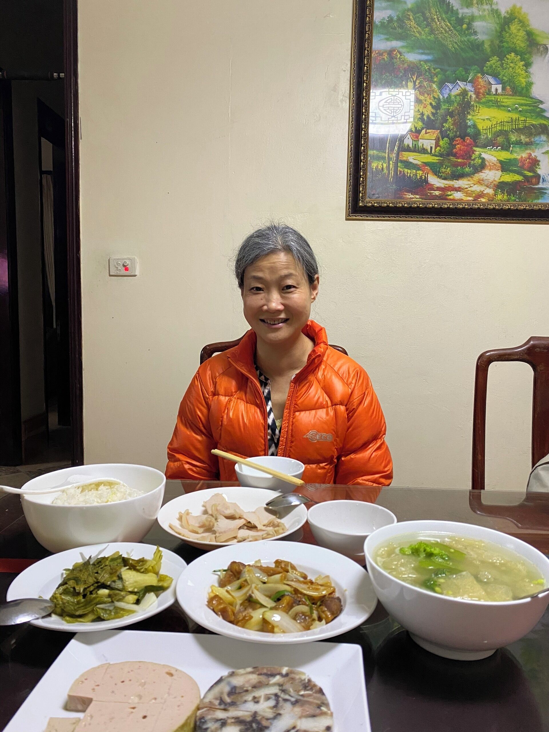 A dining table with various dishes, a person in an orange jacket, and a painting on the wall.