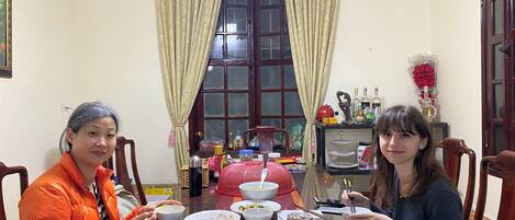 A dining table with various dishes and utensils, a window with curtains, and a small shelf with bottles and a vase.
