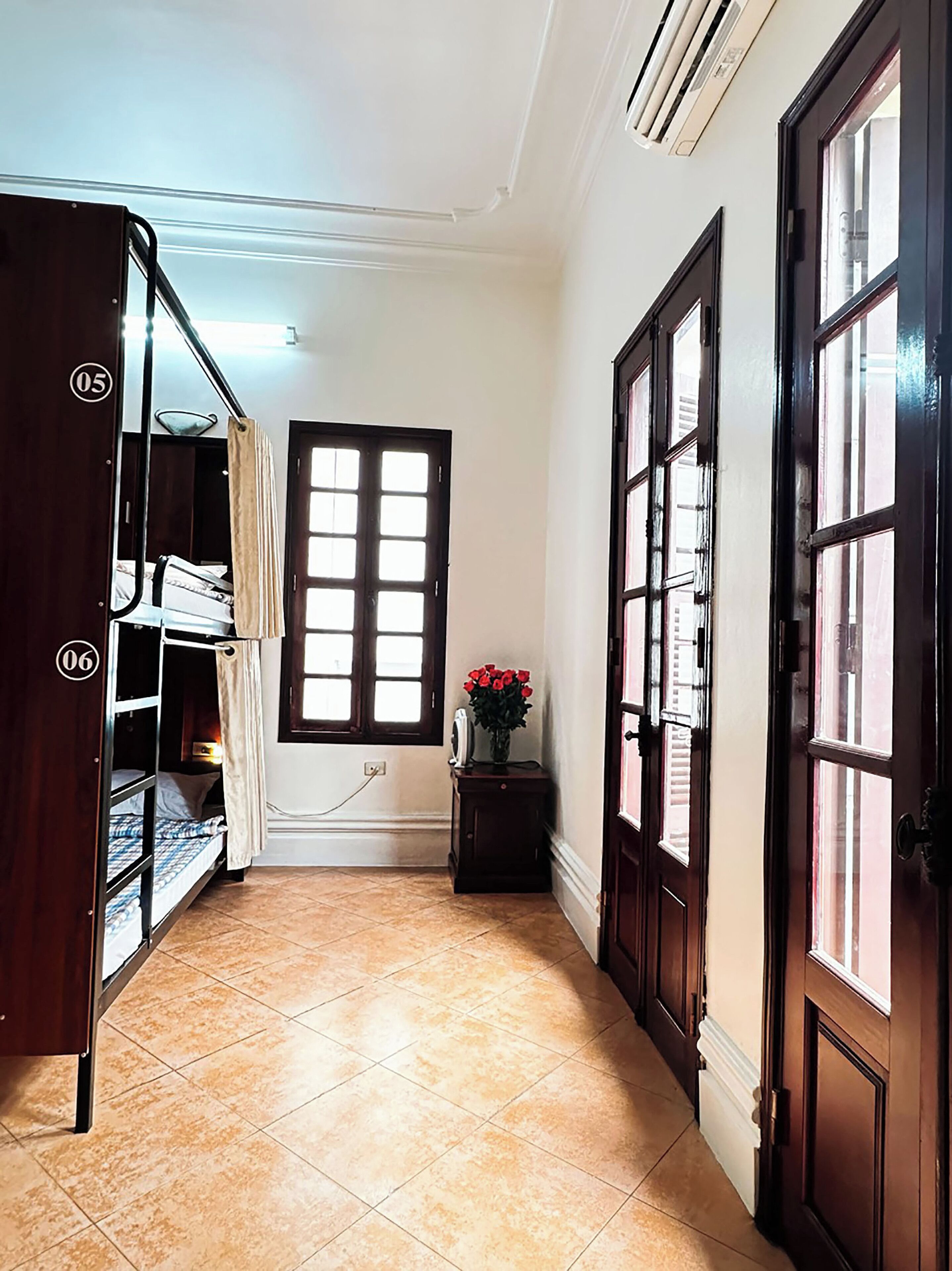 A corridor with a tiled floor, a window with blinds, a bedside table with a vase of flowers, and a door with a number plaque.
