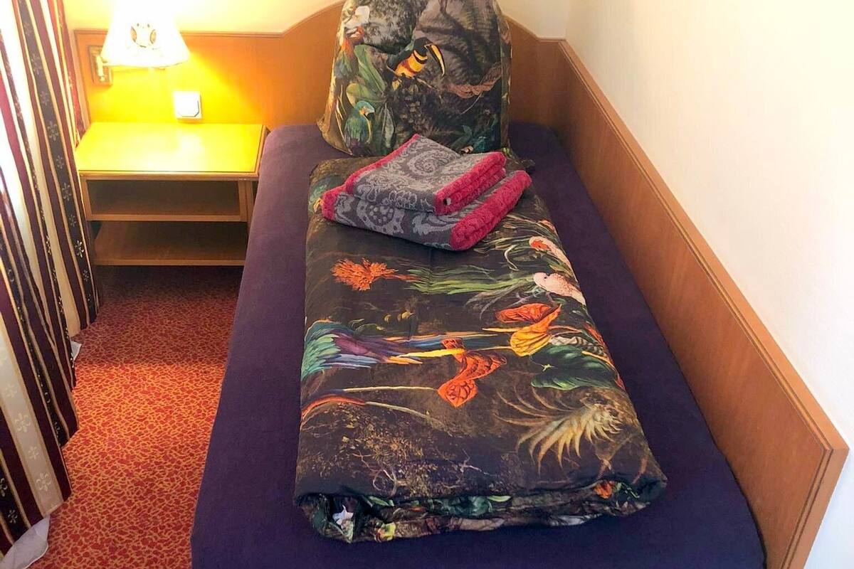 Una habitación pequeña con una cama individual, un colchón morado, una manta estampada con flores y una almohada con un diseño de pájaro.