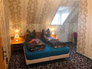 Una habitación pequeña y acogedora con techo inclinado, dos camas con ropa de cama azul, papel tapiz estampado y un ventanal que deja entrar luz natural.