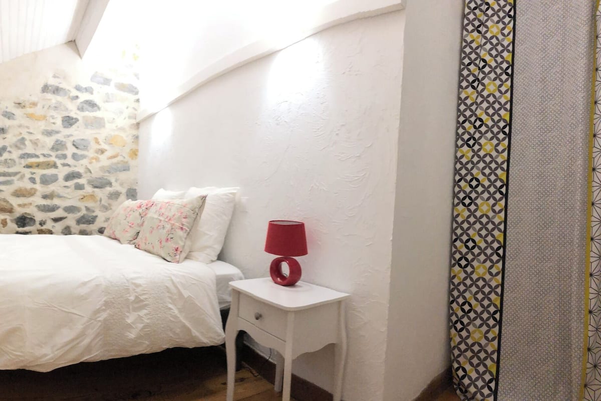 Un dormitorio con pared de piedra, una cama con ropa de cama blanca, una mesita de noche pequeña con una lámpara roja y una ventana con marco de madera.