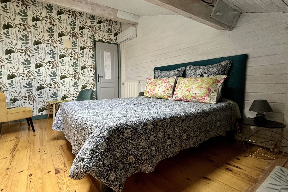 Un dormitorio con colcha estampada, cabecera verde, mesita de noche con lámpara y una silla.