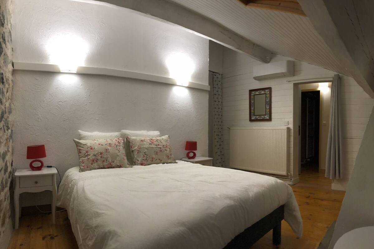 Un dormitorio con cama, mesitas de noche, una silla y un espejo en la pared.