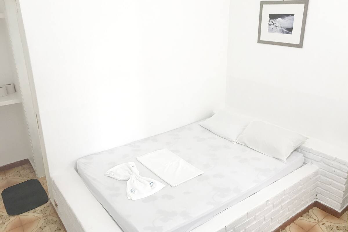 A small, white, single-bed room with a grey mattress, a white pillow, and a towel on the bed. There is a framed picture on the wall above the bed.