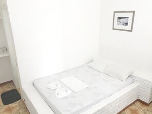 A small, white, single-bed room with a grey mattress, a white pillow, and a towel on the bed. There is a framed picture on the wall above the bed.