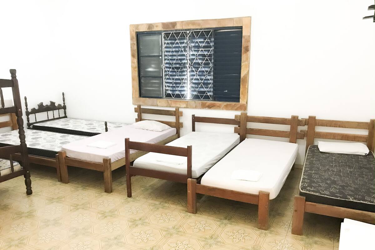A room with four wooden beds arranged in a row, a window with a metal grid, and patterned floor tiles.