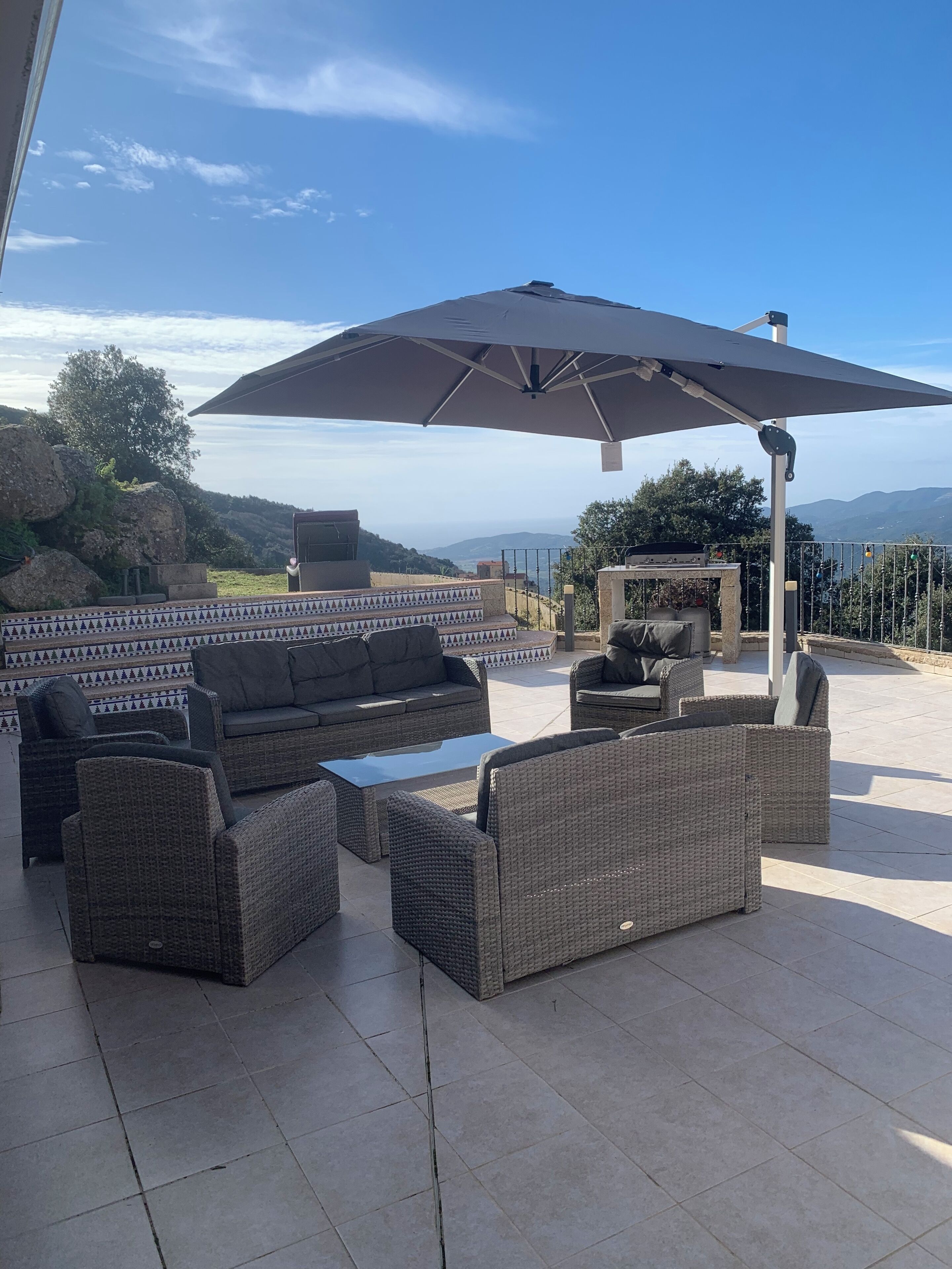 Eine Terrasse im Freien mit Korbmöbeln, einem großen Sonnenschirm und einem Steinkamin.