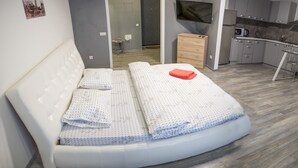 Ett modernt sovrum med en stor säng, en tv, en byrå i trä och en liten köksdel.