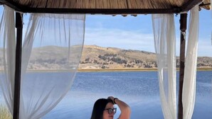Una cama con vista a un lago y colinas, un dosel arriba y una persona sentada en la cama.